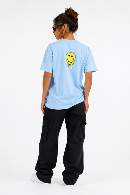 A woman shown from the back wearing a light blue t-shirt with a yellow dripping smiley face graphic on the upper back, paired with black cargo pants and white sneakers, against a plain white studio background.