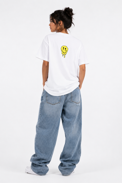 A model viewed from behind wearing a white t-shirt with a small yellow smiley face graphic on the back, paired with loose-fit light blue jeans and white sneakers, against a plain white studio background.