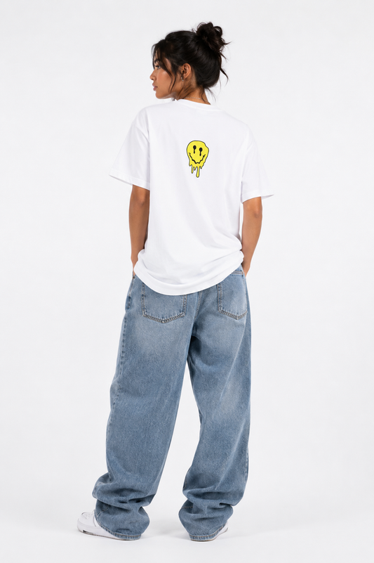 A model viewed from behind wearing a white t-shirt with a small yellow smiley face graphic on the back, paired with loose-fit light blue jeans and white sneakers, against a plain white studio background.