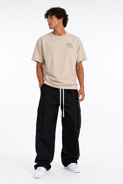 A man modeling a beige short-sleeve t-shirt with 'HALF FACE' printed on the chest, paired with black cargo pants and white sneakers, against a plain white background.