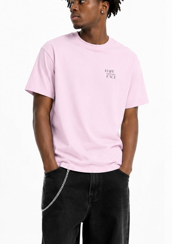 A man wearing a light pink crewneck t-shirt with a small black 'HAPPY FACE' graphic on the left chest, paired with black jeans and a silver chain belt, against a plain white background.